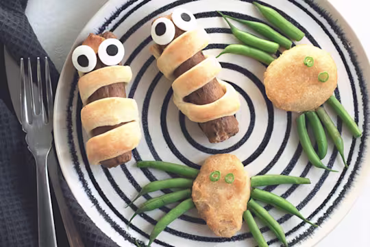 Quorn Sausages wrapped in shortcrust pastry with edible eyes and Quorn Crispy Nuggets with green bean 'legs' to resemble spiders on a black and white plate.