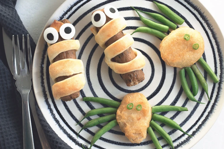 Quorn Sausages wrapped in shortcrust pastry with edible eyes and Quorn Crispy Nuggets with green bean 'legs' to resemble spiders on a black and white plate.