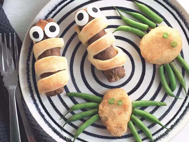 Quorn Sausages wrapped in shortcrust pastry with edible eyes and Quorn Crispy Nuggets with green bean 'legs' to resemble spiders on a black and white plate.