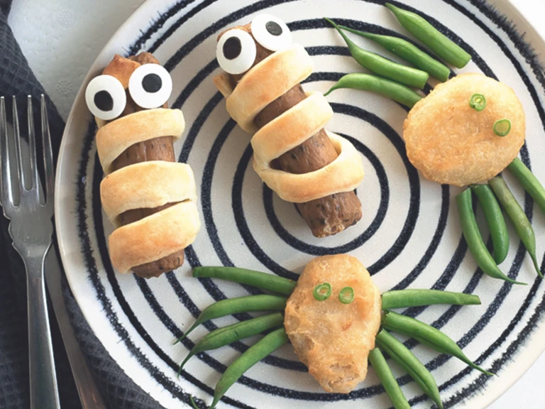 Quorn Sausages wrapped in shortcrust pastry with edible eyes and Quorn Crispy Nuggets with green bean 'legs' to resemble spiders on a black and white plate.