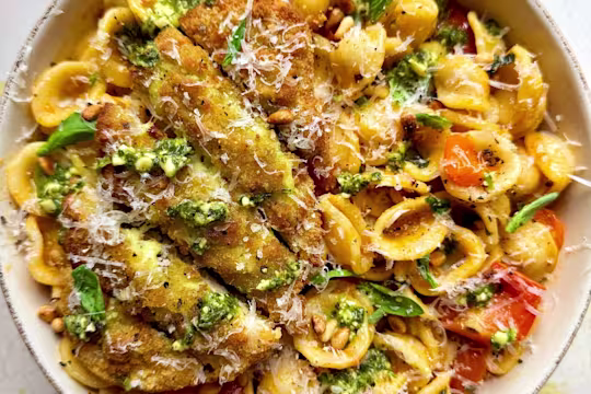A bowl of Tomato Orecchiette Pasta with Quorn Mozzarella and Pesto Escalopes, topped with garlic, cherry tomatoes, chilli flakes, basil, parmesan and pine nuts on a white background.