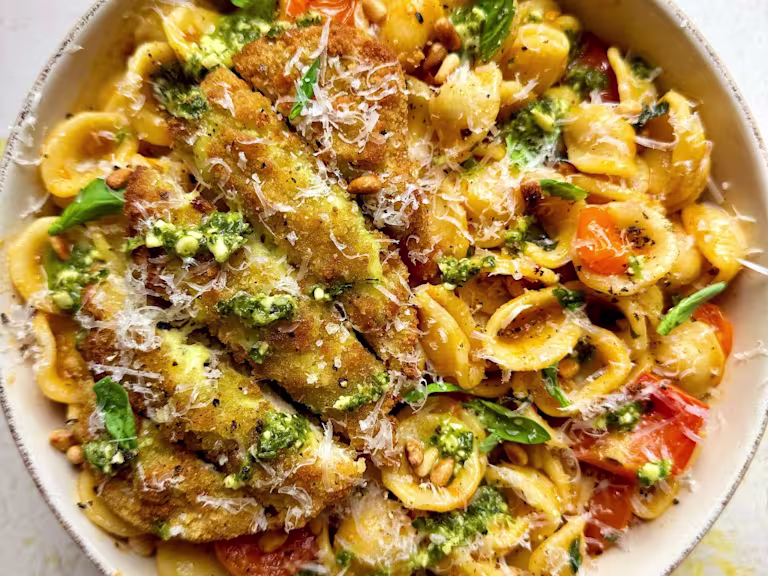 A bowl of Tomato Orecchiette Pasta with Quorn Mozzarella and Pesto Escalopes, topped with garlic, cherry tomatoes, chilli flakes, basil, parmesan and pine nuts on a white background.