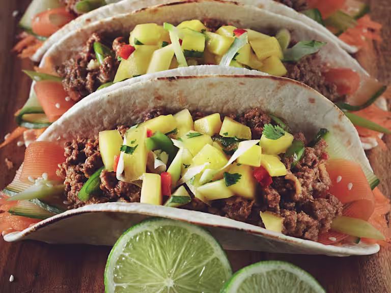 Two tacos in flour tortillas filled with ribbons of carrot and zucchini and Quorn Meatless Grounds, topped with mango salsa and served on a wooden plank with a wedge of lime on the side.