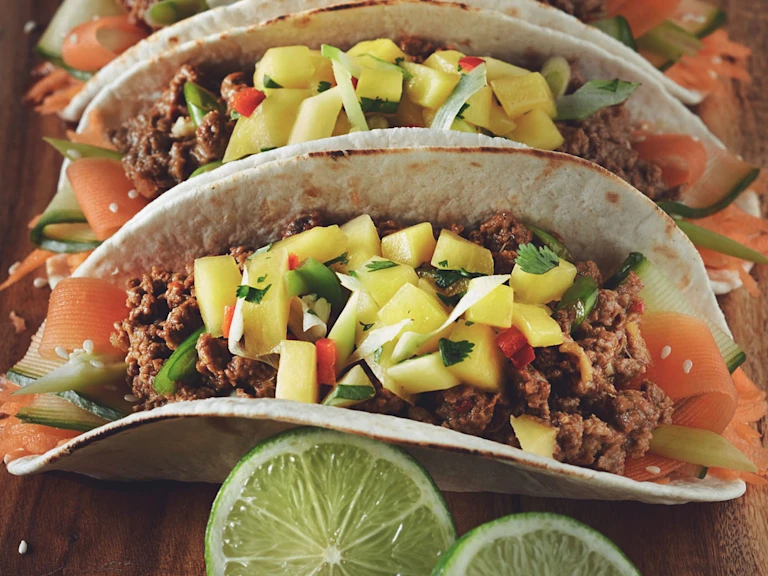 Two tacos in flour tortillas filled with ribbons of carrot and zucchini and Quorn Meatless Grounds, topped with mango salsa and served on a wooden plank with a wedge of lime on the side.