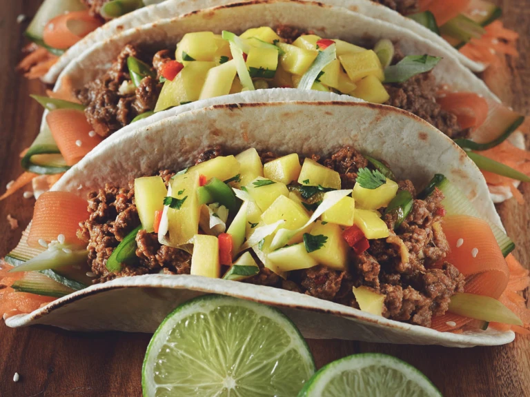 Two tacos in flour tortillas filled with ribbons of carrot and zucchini and Quorn Meatless Grounds, topped with mango salsa and served on a wooden plank with a wedge of lime on the side.