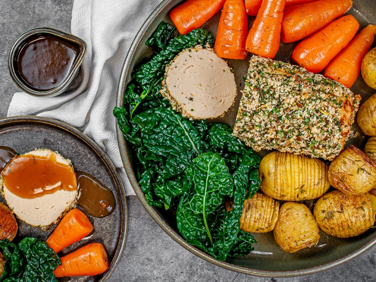 Herb crusted Quorn roast sliced served alongside potatoes, carrots and greens with a pot of gravy on the side.