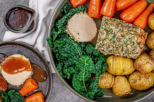 Herb crusted Quorn roast sliced served alongside potatoes, carrots and greens with a pot of gravy on the side.