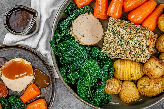 Herb crusted Quorn roast sliced served alongside potatoes, carrots and greens with a pot of gravy on the side.