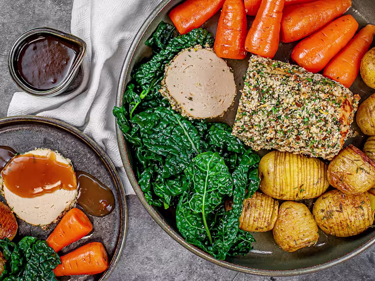 Herb crusted Quorn roast sliced served alongside potatoes, carrots and greens with a pot of gravy on the side.