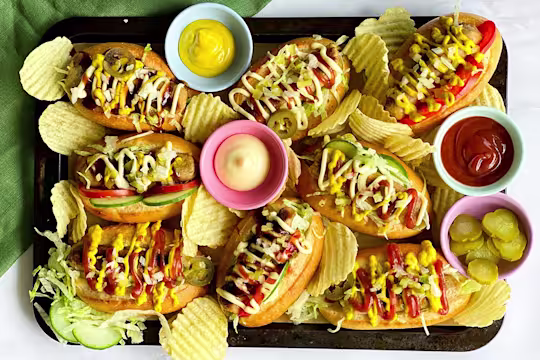A platter of Chicago-style hot dogs made with Quorn sausages on bun topped with onion, dill pickles, tomatoes, cucumber, jalapenos, and lettuce with sauces and crisps on the side.