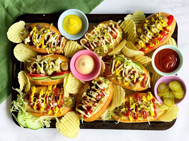 A platter of Chicago-style hot dogs made with Quorn sausages on bun topped with onion, dill pickles, tomatoes, cucumber, jalapenos, and lettuce with sauces and crisps on the side.