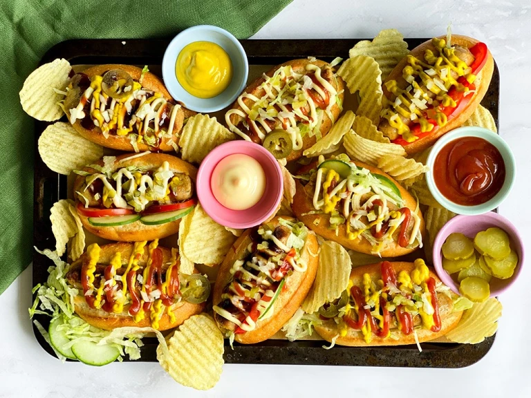 A platter of Chicago-style hot dogs made with Quorn sausages on bun topped with onion, dill pickles, tomatoes, cucumber, jalapenos, and lettuce with sauces and crisps on the side.