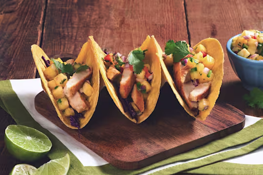 Three hard-shell corn tacos filled with Quorn Fillets, red cabbage, and pineapple salsa arranged on a wooden board with half of a lime and a blue ramekin of pineapple salsa on the side.