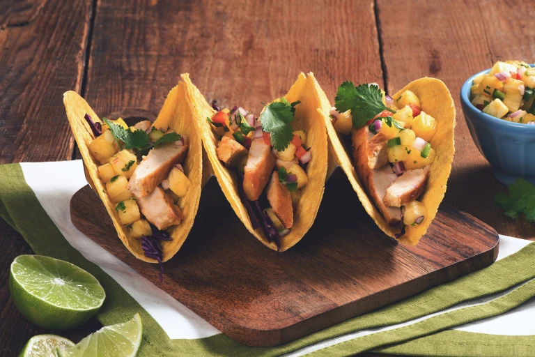 Three hard-shell corn tacos filled with Quorn Fillets, red cabbage, and pineapple salsa arranged on a wooden board with half of a lime and a blue ramekin of pineapple salsa on the side.