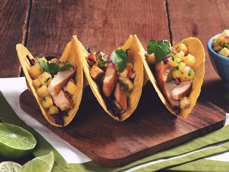Three hard-shell corn tacos filled with Quorn Fillets, red cabbage, and pineapple salsa arranged on a wooden board with half of a lime and a blue ramekin of pineapple salsa on the side.
