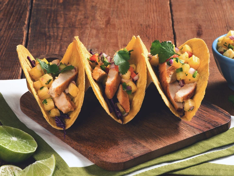 Three hard-shell corn tacos filled with Quorn Fillets, red cabbage, and pineapple salsa arranged on a wooden board with half of a lime and a blue ramekin of pineapple salsa on the side.