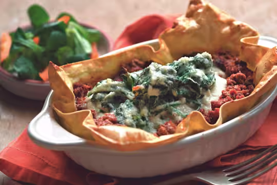 Baked sheets of lasagna topped with Quorn Grounds and a ricotta basil topping in a white oven dish with a salad in the background.