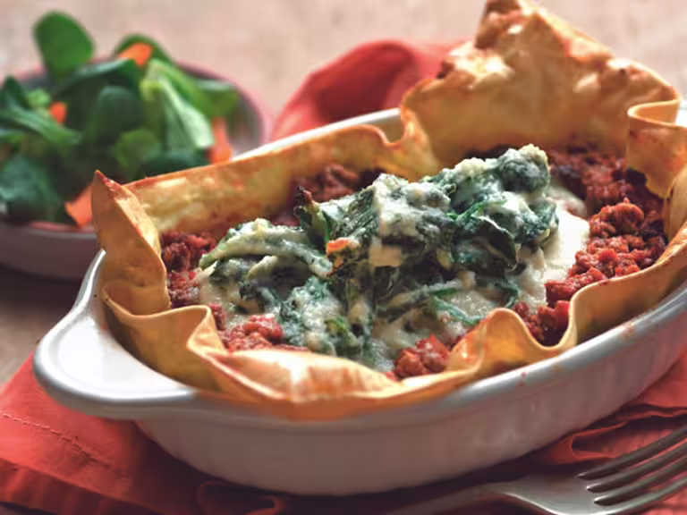 Baked sheets of lasagna topped with Quorn Grounds and a ricotta basil topping in a white oven dish with a salad in the background.