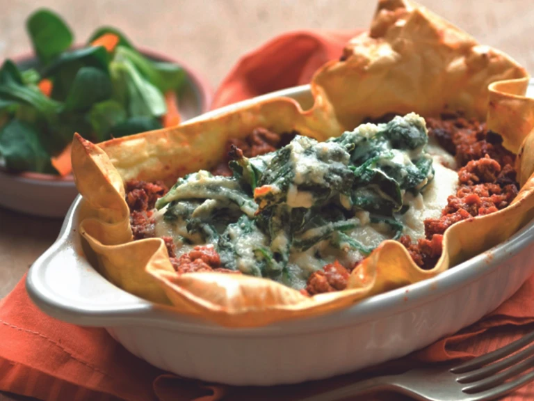 Baked sheets of lasagna topped with Quorn Grounds and a ricotta basil topping in a white oven dish with a salad in the background.