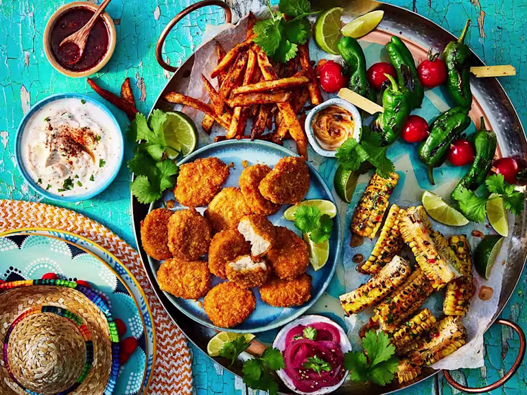 Quorn Cheesy Nacho Nuggs on a sharing platter with corn on the cob, chips, and tomato and lettuce skewers. Small sombrero and dips to the side.