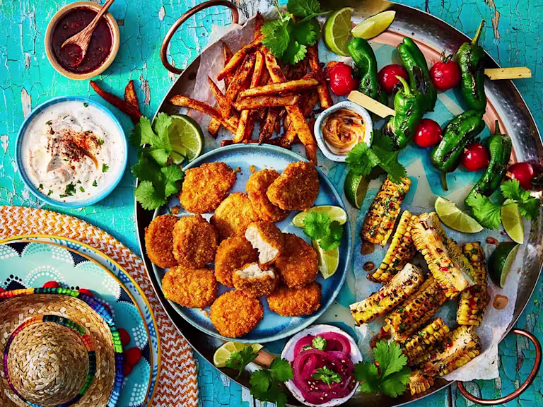 Quorn Cheesy Nacho Nuggs on a sharing platter with corn on the cob, chips, and tomato and lettuce skewers. Small sombrero and dips to the side.