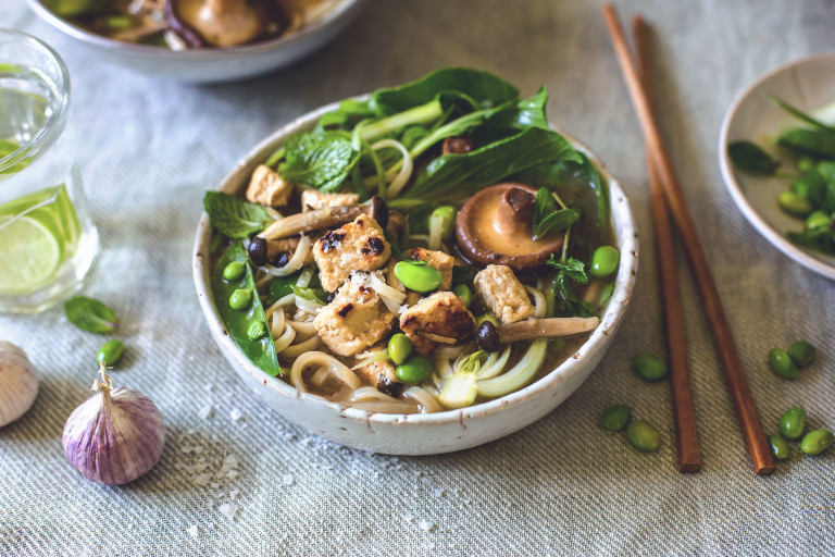 A bowl of rice noodle soup topped with  mushrooms, peas, edamame, fresh herbs, bok choy, and Quorn Pieces, with a scattering of salt and edamame, a head of purple garlic, and chopsticks surrounding it.
