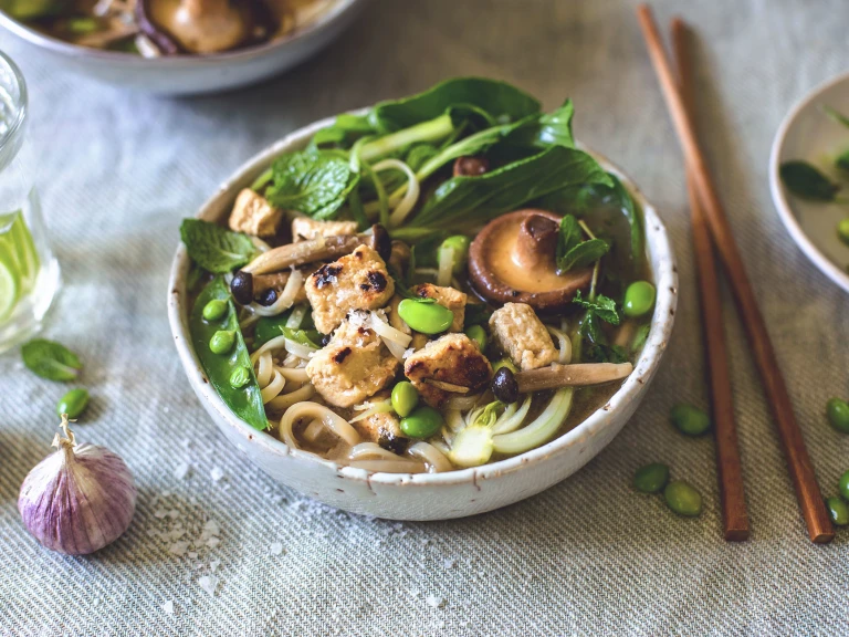 A bowl of rice noodle soup topped with  mushrooms, peas, edamame, fresh herbs, bok choy, and Quorn Pieces, with a scattering of salt and edamame, a head of purple garlic, and chopsticks surrounding it.