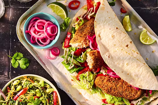 Quorn Salt and Pepper Tenders in a tortilla wrap with lettuce, chopped pepper, pickled red onion. Lime wedges are on the serving tray and a bowl of Chinese chopped slaw to the side.