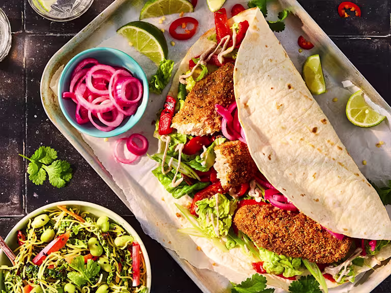 Quorn Salt and Pepper Tenders in a tortilla wrap with lettuce, chopped pepper, pickled red onion. Lime wedges are on the serving tray and a bowl of Chinese chopped slaw to the side.