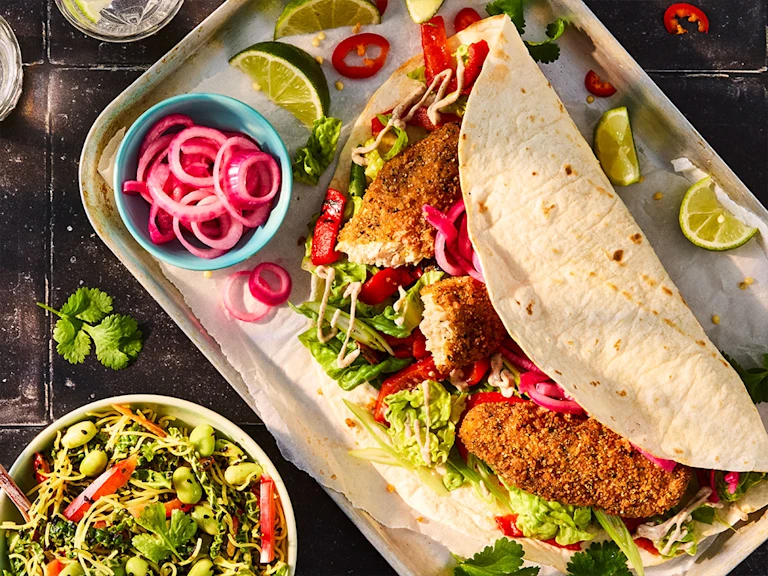 Quorn Salt and Pepper Tenders in a tortilla wrap with lettuce, chopped pepper, pickled red onion. Lime wedges are on the serving tray and a bowl of Chinese chopped slaw to the side.