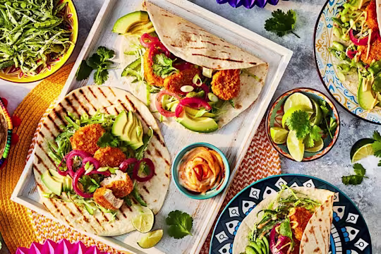 Quorn Cheesy Nacho Nuggs in open chargrilled tortillas with salad, chopped limes to garnish and bowls of salad to the side.