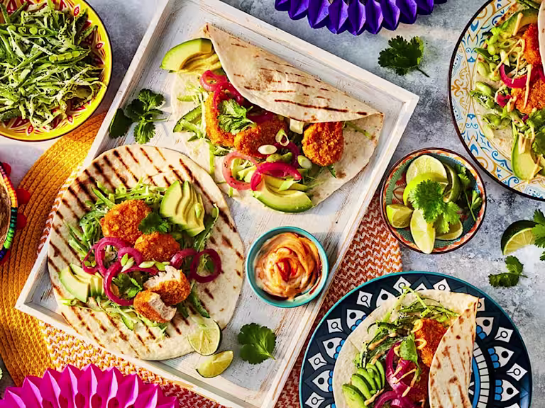 Quorn Cheesy Nacho Nuggs in open chargrilled tortillas with salad, chopped limes to garnish and bowls of salad to the side.