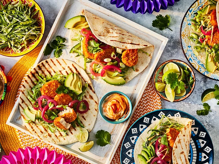 Quorn Cheesy Nacho Nuggs in open chargrilled tortillas with salad, chopped limes to garnish and bowls of salad to the side.