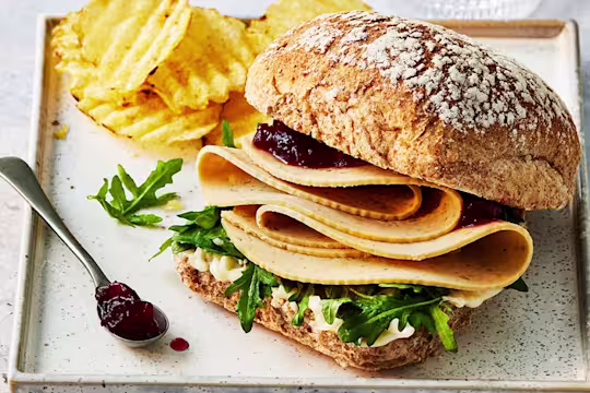 Quorn turkey slices in a sandwich with rocket leaves and cranberry sauce with crisps on a tray