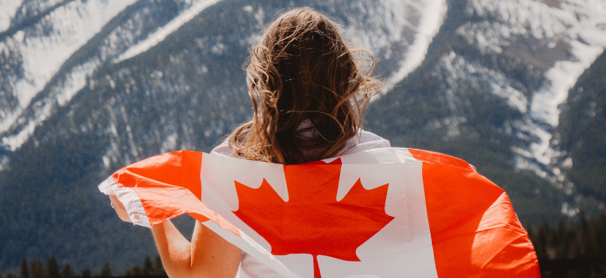 A woman holds a Canadian flag around her back as she looks at a mountain range.