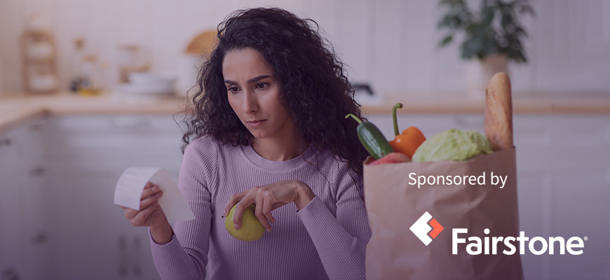 A women sits at a kitchen table with a bag of groceries next to her. She is looking concerned as she reviews a receipt.