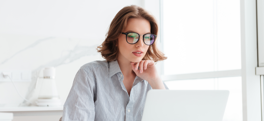 A young woman sits at a table, looking at a laptop computer.