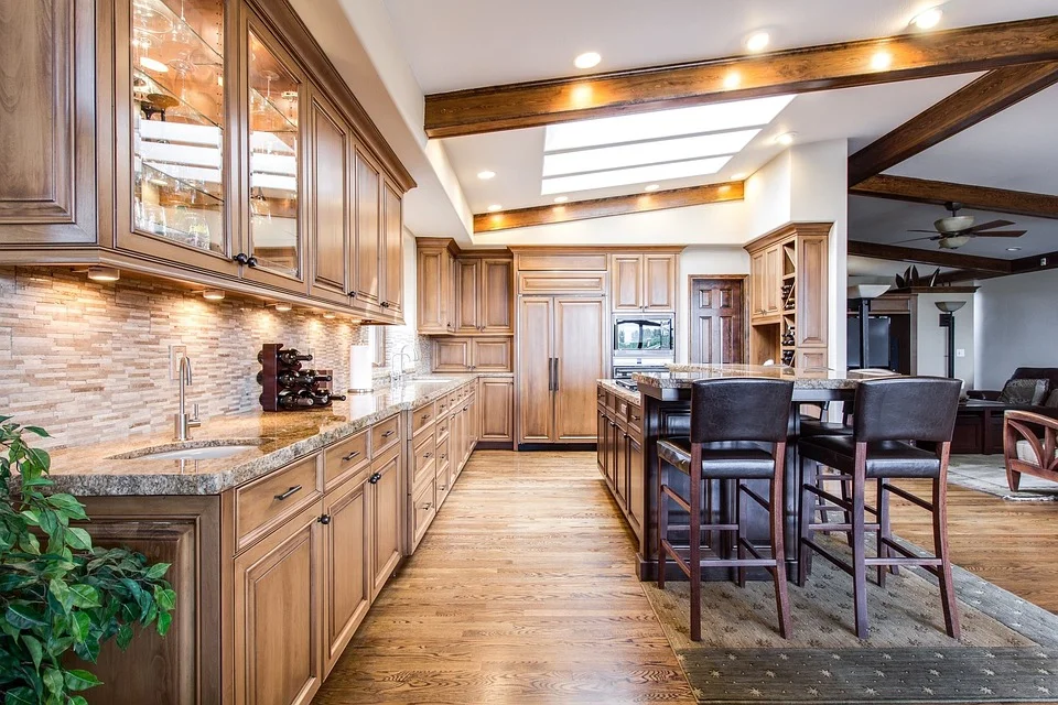 Spacious rustic kitchen featuring light wood cabinetry, granite countertops, brick backsplash, exposed wooden ceiling beams with recessed lighting, stainless steel appliances, island with black bar stools, and hardwood flooring throughout the open-concept space.
