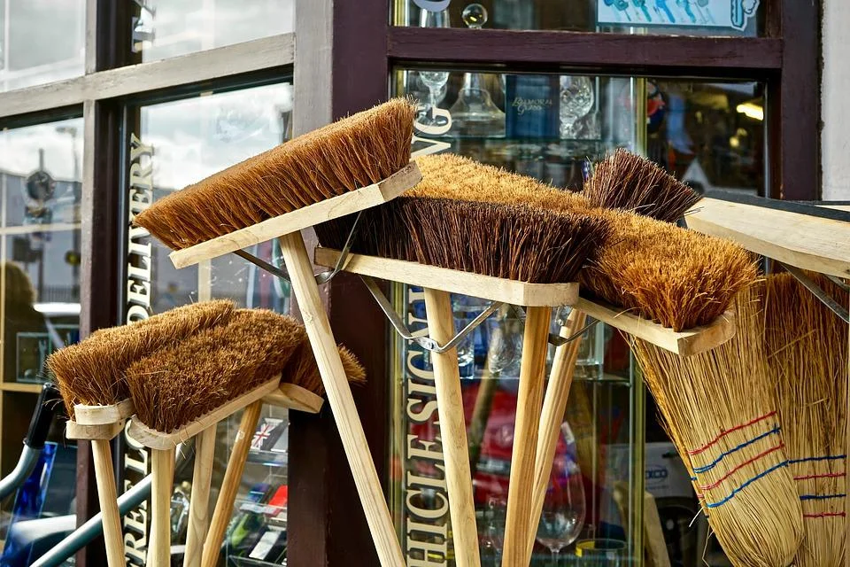 Multiple brooms with natural brown and tan bristles attached to wooden handles, arranged in a row against a storefront window displaying merchandise in the background.