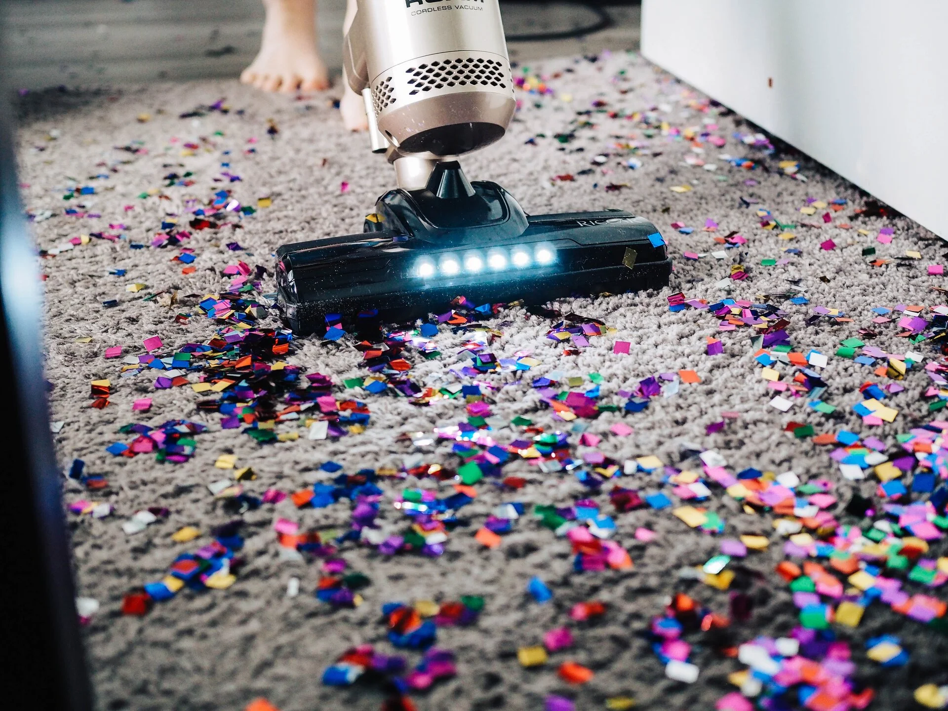 White cordless vacuum with illuminated brush head cleaning gray carpet covered in colorful confetti pieces in various bright colors including pink, blue, purple, and yellow.
