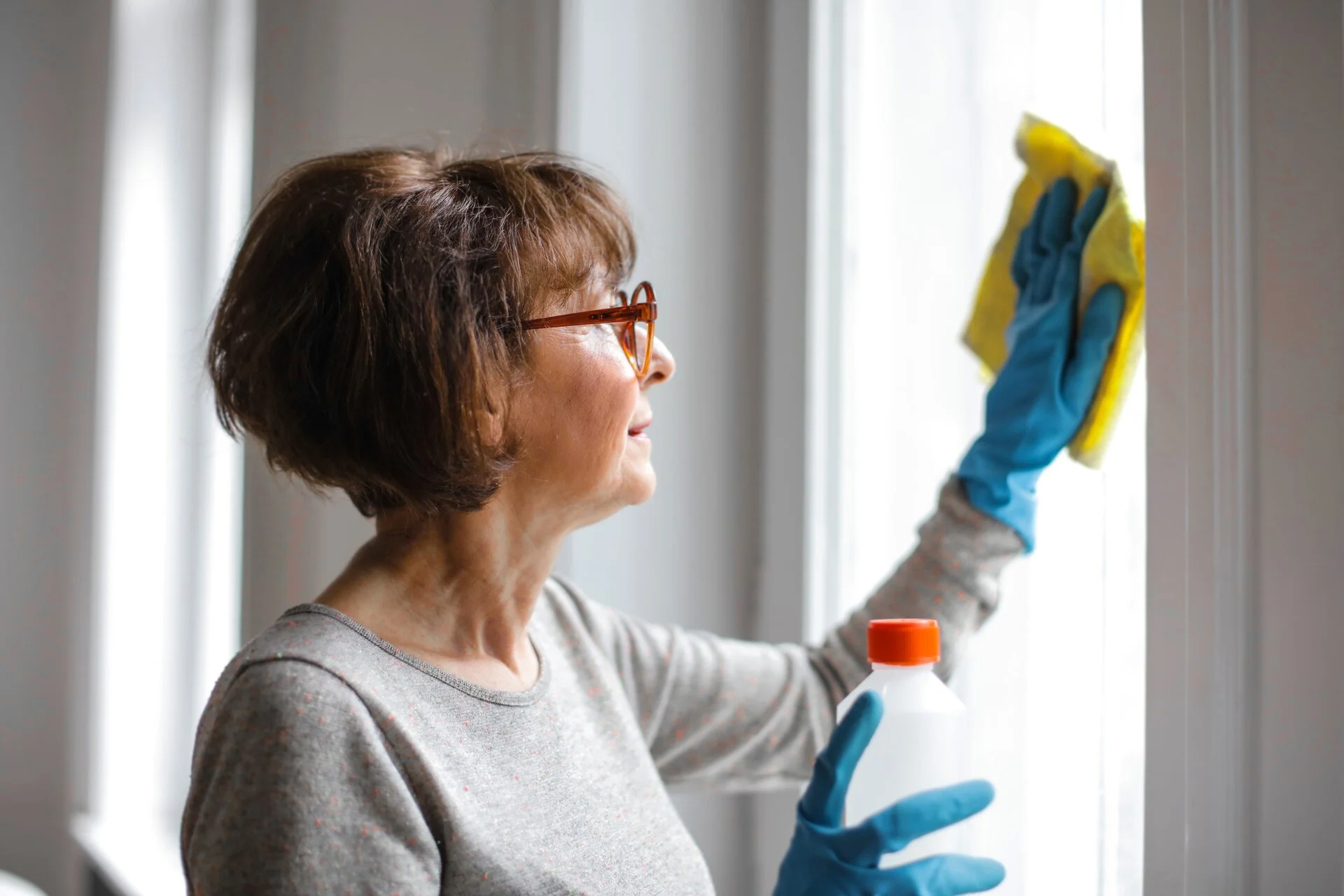 Middle-aged woman wearing brown glasses and gray shirt uses a yellow squeegee with blue cloth to clean a bright window, wearing blue rubber gloves.