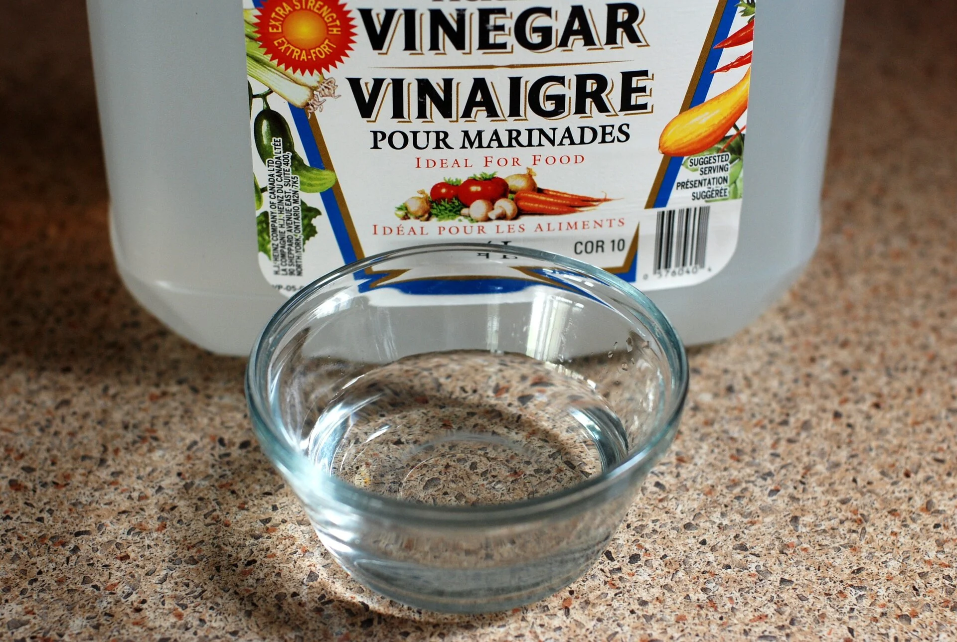 Clear glass bowl containing vinegar solution on speckled countertop, with white plastic vinegar bottle labeled 'Pour Marinades' and bilingual text behind it.