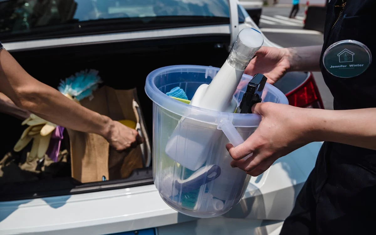 Two people holding a clear plastic bucket filled with cleaning supplies including a white spray bottle, blue brush, and black cleaning tools, with a car and Jennifer Winter branding visible in background