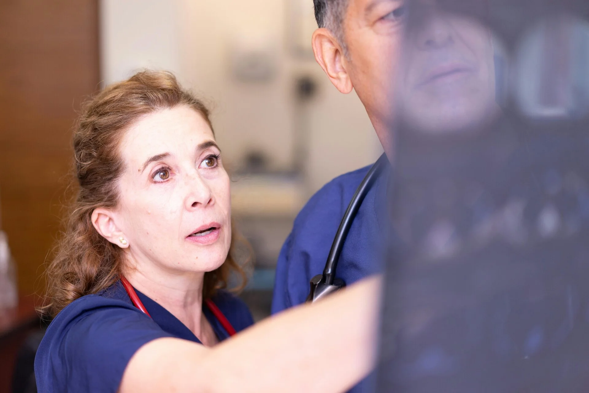 Female healthcare worker in blue scrubs with stethoscope engaged in conversation with male colleague in clinical setting, both appearing focused and attentive during discussion.