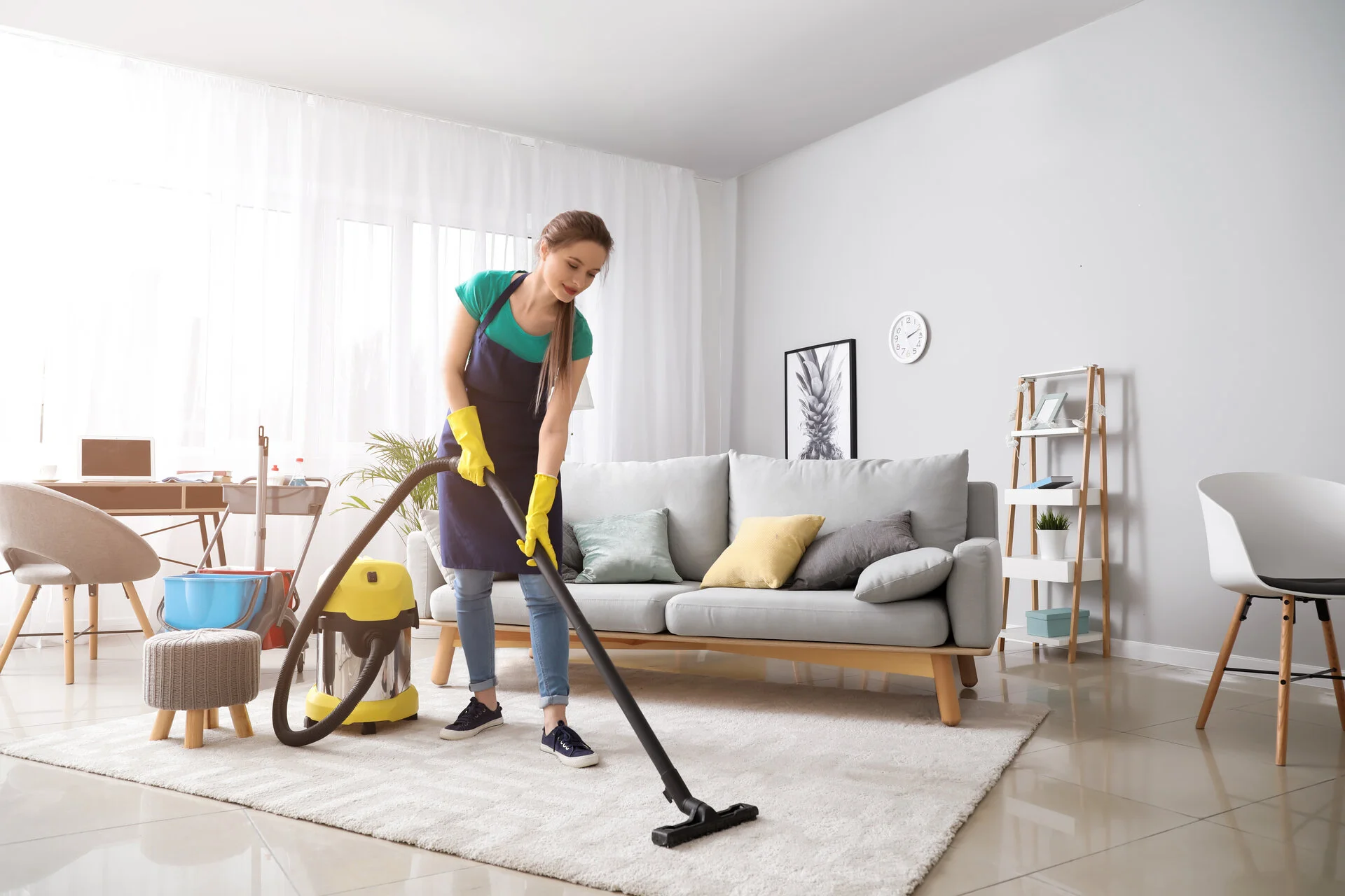 Woman in teal shirt and yellow gloves uses a yellow and black vacuum cleaner on a light beige carpet in a bright, modern living room with gray sofa and white walls.