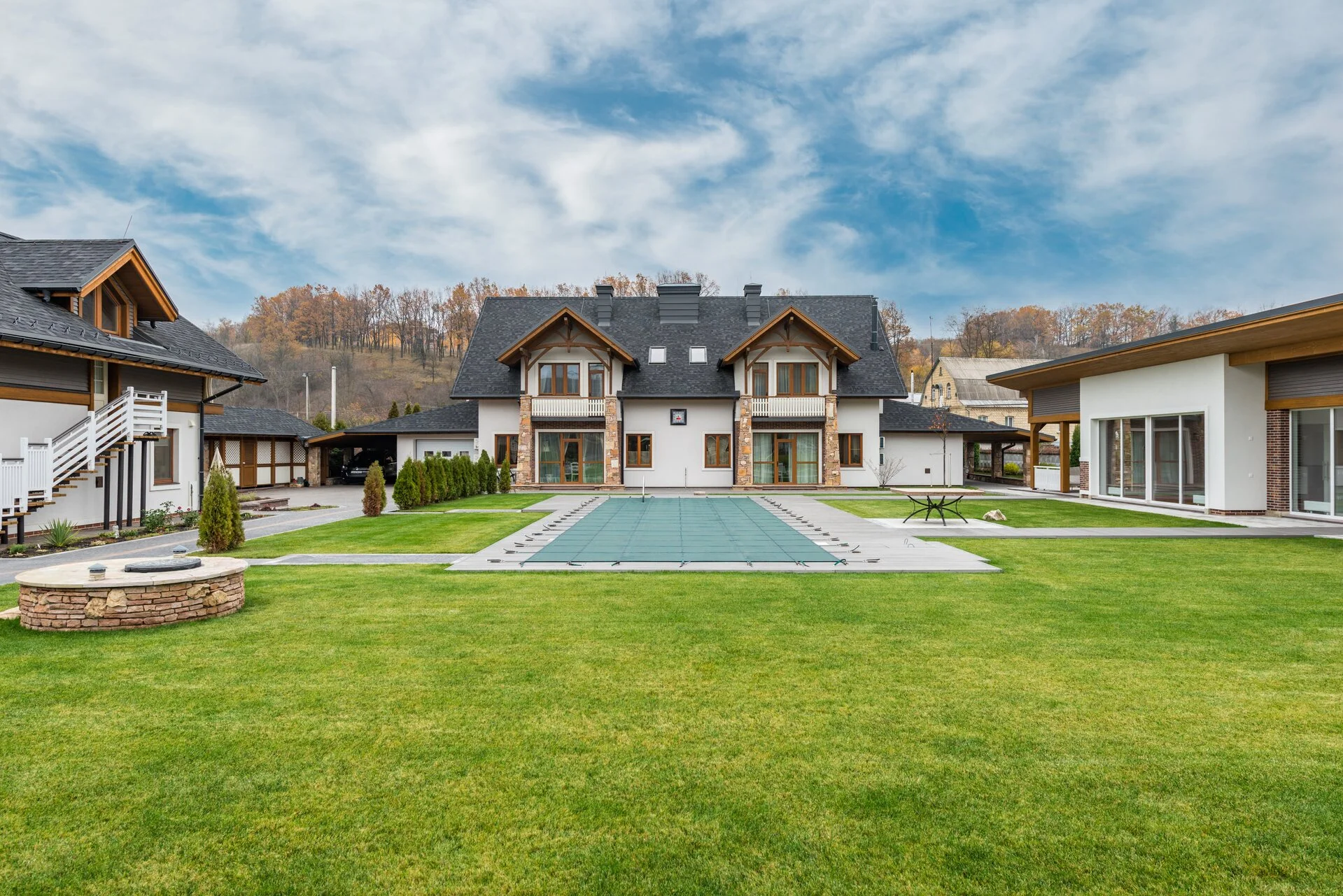 Expansive green lawn in a residential courtyard surrounded by contemporary white and stone houses with covered pool, fire pit, and autumn trees under blue sky with white clouds
