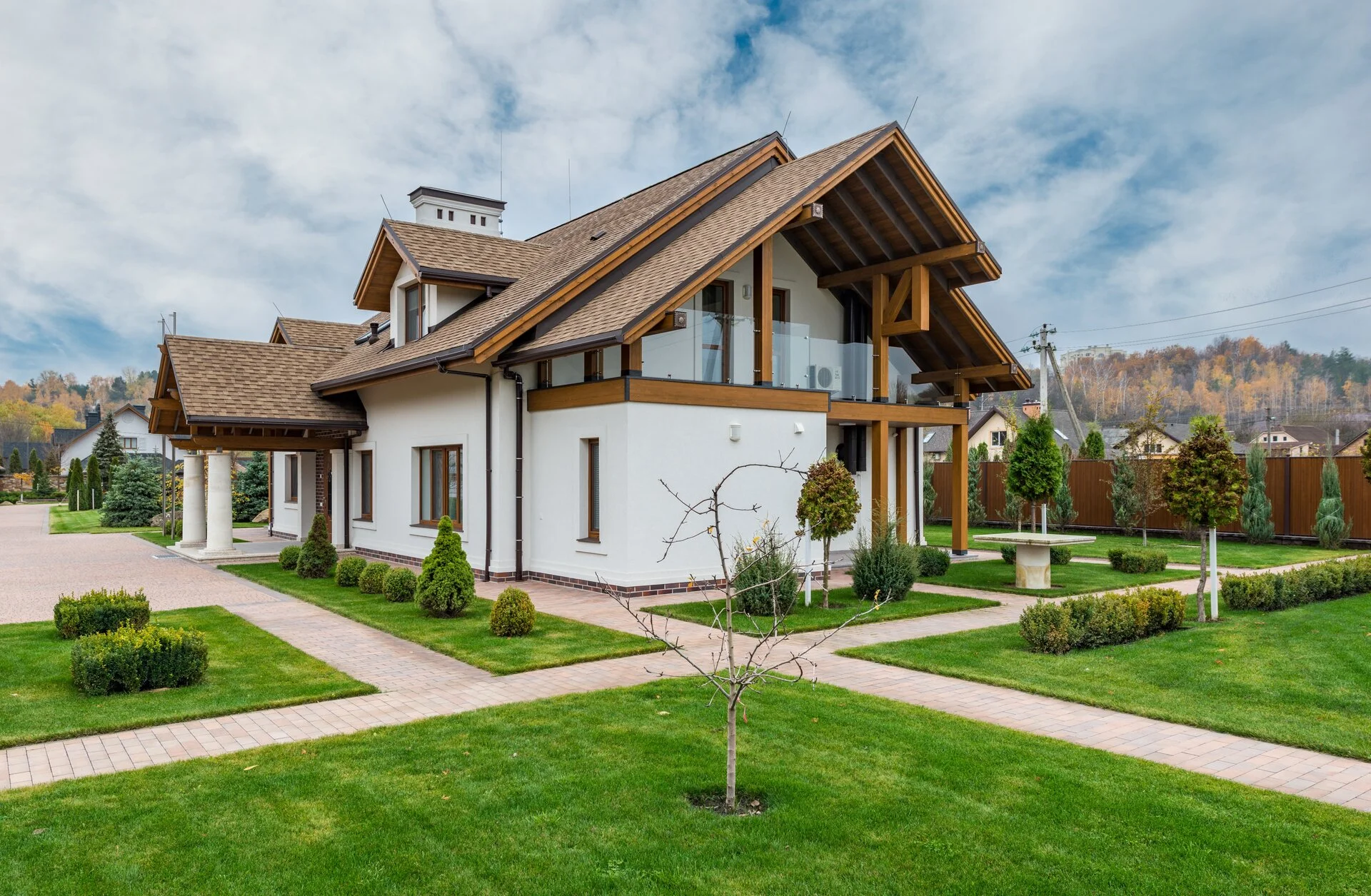 Contemporary white house with brown wooden trim and gabled roofs, featuring a manicured green lawn, brick pathway, ornamental shrubs, and forested hillside backdrop under partly cloudy sky.
