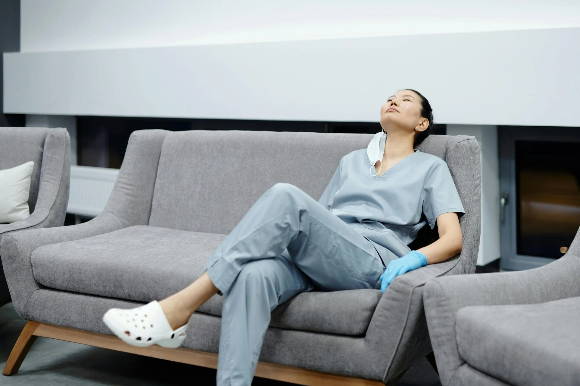 A female healthcare professional wearing light blue scrubs and blue latex gloves sits on a gray upholstered couch, looking upward with a relaxed expression. She wears white clogs and has a stethoscope around her neck in a modern medical facility setting.
