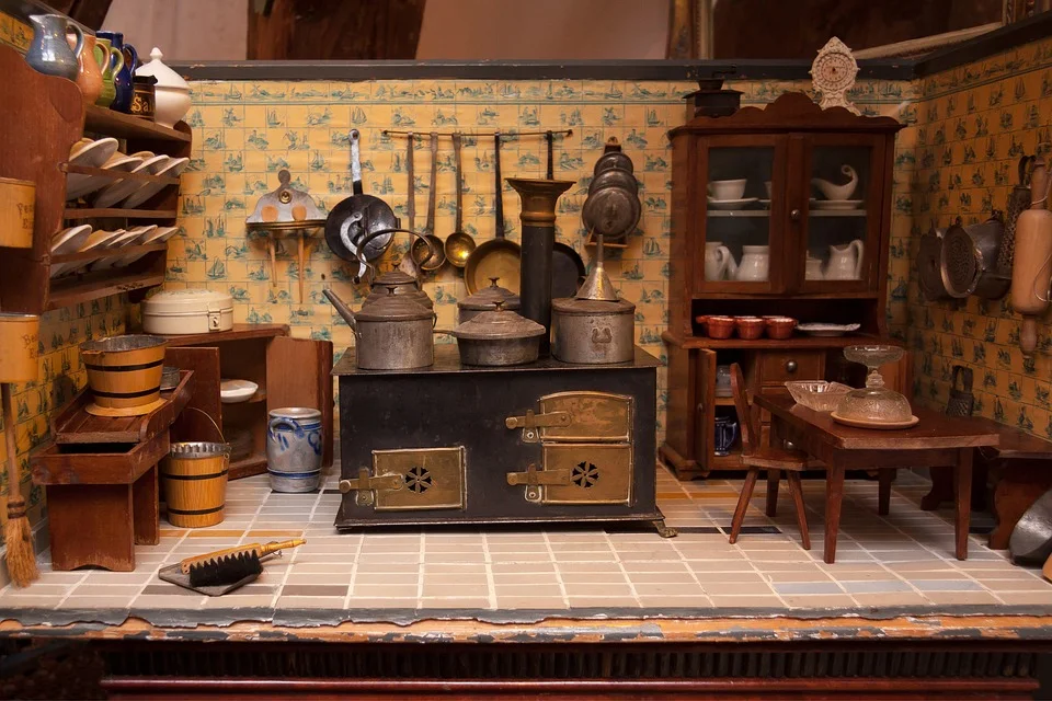 Detailed dollhouse kitchen featuring a black cast iron stove with brass accents, copper cookware hanging on yellow patterned walls, wooden furniture including a china cabinet and table, ceramic dishware, and period-appropriate accessories on a tiled floor.