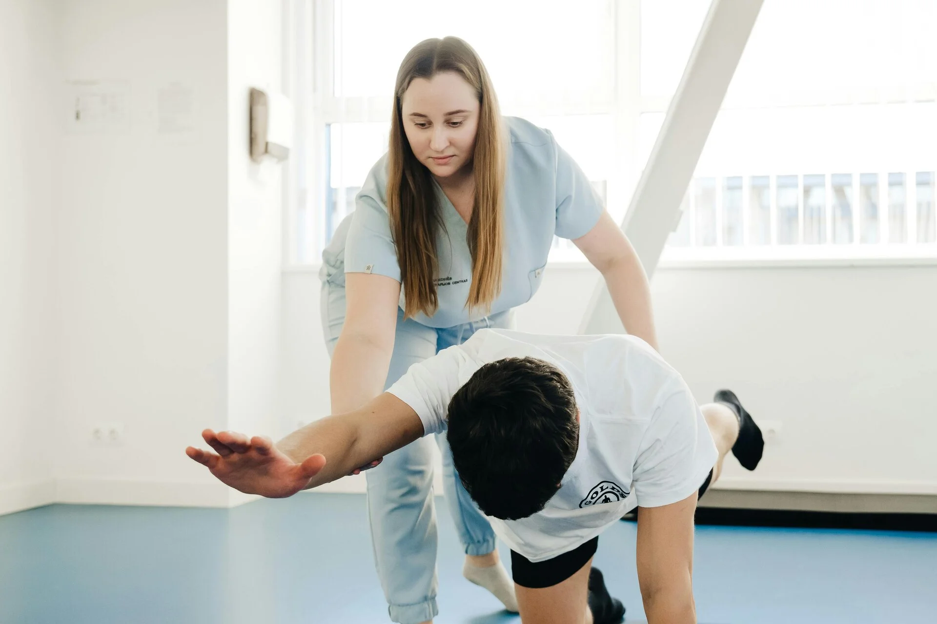 Female physiotherapist in light blue uniform guides male patient in white shirt through a forward bend stretch in a bright clinical room with blue flooring and large windows.
